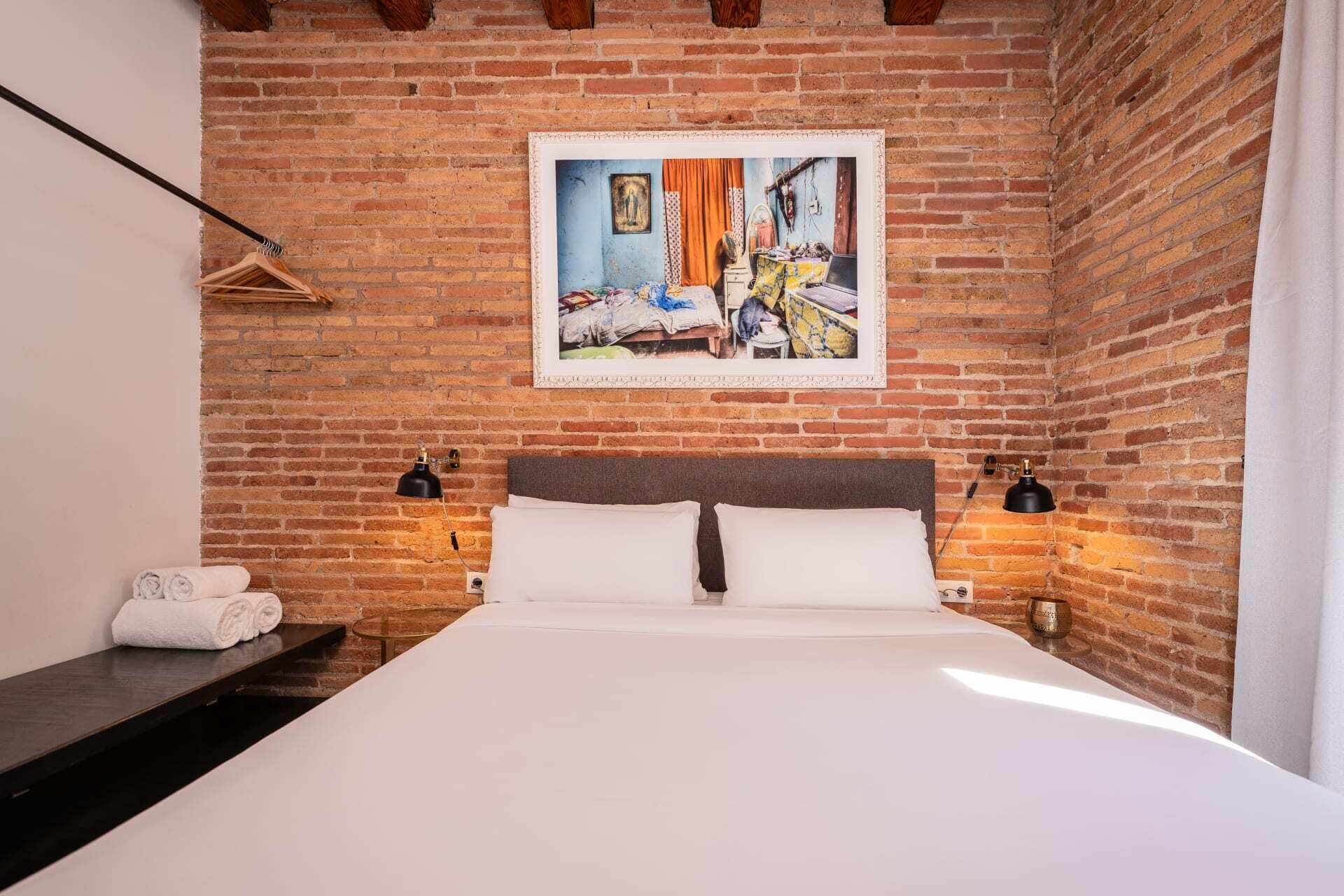 A bedroom with a brick wall, a bed with white linens, two bedside lamps, and a framed picture above the bed.