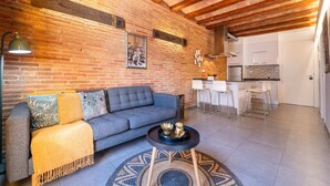 Un salon chaleureux avec un canapé bleu, une table basse ronde et un tapis à motifs. On aperçoit à l’arrière-plan une cuisine avec des meubles blancs et un coin repas équipé de tabourets de bar.