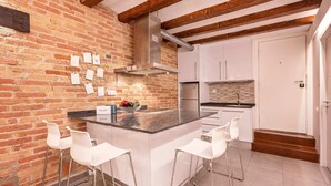 Une cuisine avec un mur en briques, des meubles blancs, un îlot central doté de tabourets et un réfrigérateur en acier inoxydable.