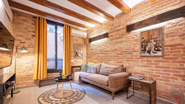 Un salon chaleureux avec des murs en briques apparentes, un plafond en bois et une grande fenêtre agrémentée de rideaux.