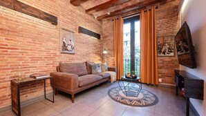 A cozy living room with exposed brick walls, a brown leather sofa, a coffee table with decorative items, and a large window with curtains.