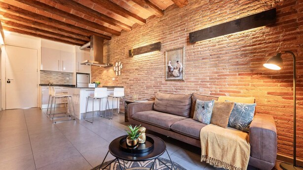 A modern kitchen with white cabinets and a brick wall. There is a brown sofa with cushions and a small round table with a plant.