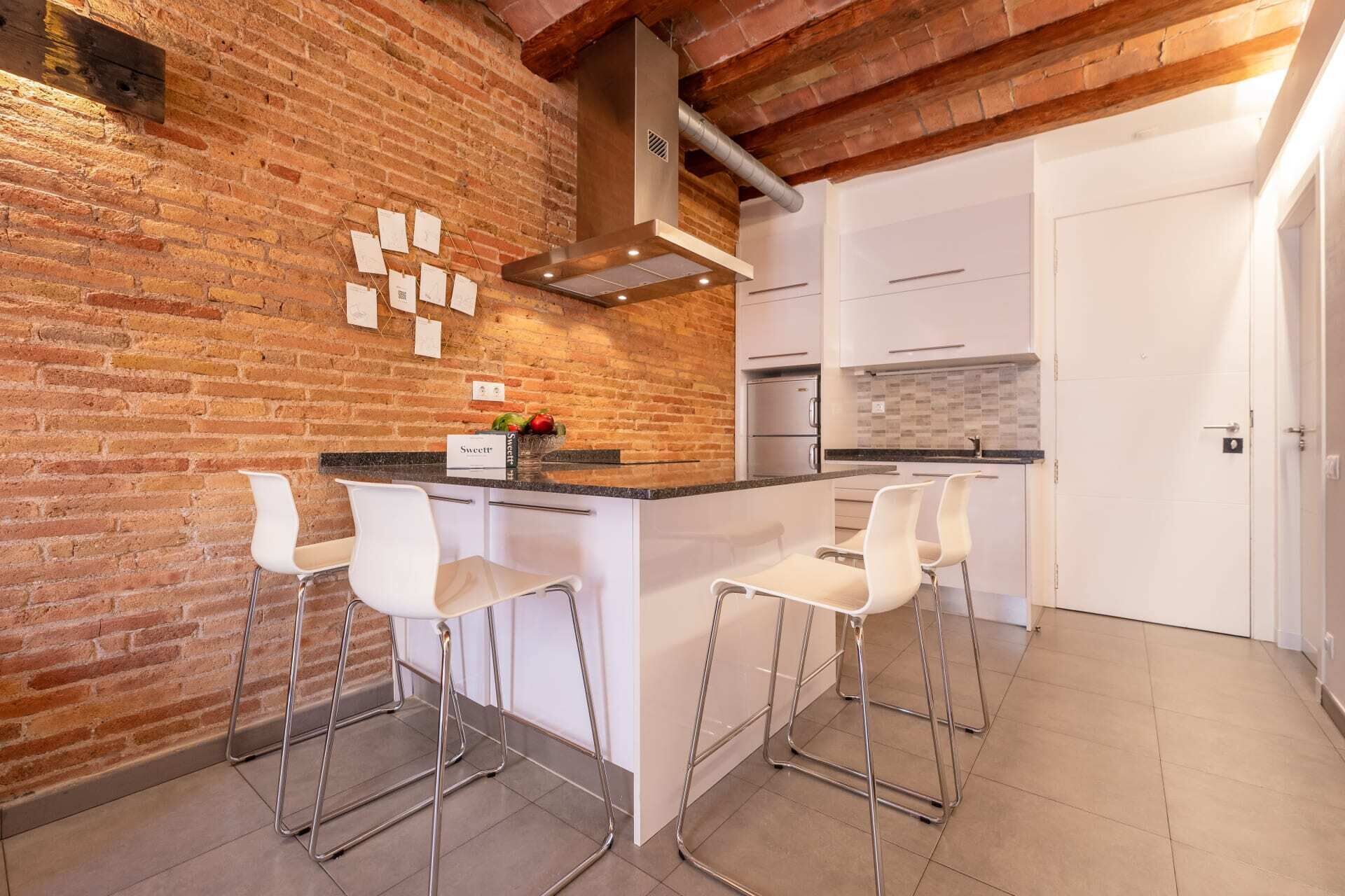 A kitchen with a brick wall, white cabinetry, a stainless steel range hood, and three white bar stools.