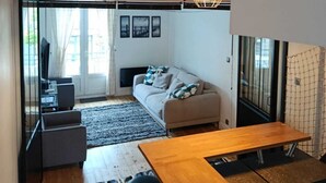 A modern living room with a sofa, a wooden dining table, and a black chair. There is a clock on the wall and a hanging light fixture. - FI Bis Saint Malo (Saint-Malo)