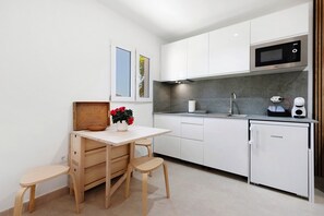 A modern kitchen with white cabinets, a microwave, a sink, and a small refrigerator. There is a wooden table with two stools and a potted plant with red flowers.