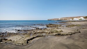 A coastal beach with rocky terrain, clear blue sky, and white buildings in the background. - Holidays 'La Lajita Beach Studio' with Sea View, Wi-Fi and Air Conditioning (La Lajita)