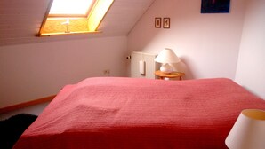 A bedroom with a skylight, a bed with a red cover, a bedside table with a lamp, and a radiator.