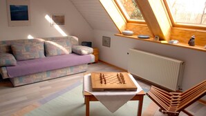 A cozy room with a loft bed, a wooden table with a chessboard, a sofa, and a window with a view.