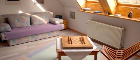 A cozy room with a loft bed, a wooden table with a chessboard, a sofa, and a window with a view.