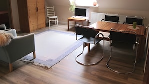 Een kamer met een schuine kap, een houten eettafel, stoelen, een klein bureau, een stoel, een kast en een raam met uitzicht op groen.
