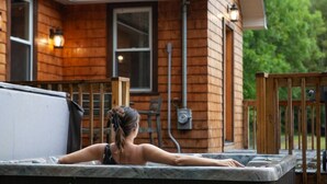 Une personne se détend dans un bain à remous à l’extérieur d’une maison en bois dotée d’une terrasse et de mobilier d’extérieur.