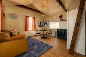 A living room with a brown leather sofa, a blue rug, a dining table with chairs, a TV, and a kitchen area with a stove. - Catskill Tiny Home, King Bed & Hot Tub on 18 acres (Ellenville)