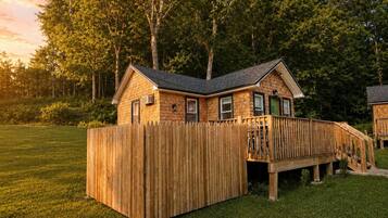 A wooden cabin with a deck, surrounded by a grassy yard and trees.
