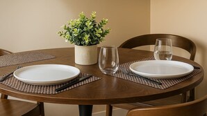 Une table de salle à manger ronde, avec deux assiettes blanches, des couverts et un verre transparent. Un set de table en fibres tressées est placé sous chaque assiette, et une plante en pot se trouve sur la table.