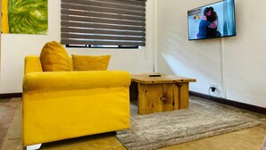 A living room with a yellow sofa, a wooden coffee table, a television mounted on the wall, and a window with blinds. - Amaryllis Residences Mount Pleasant (Blantyre)