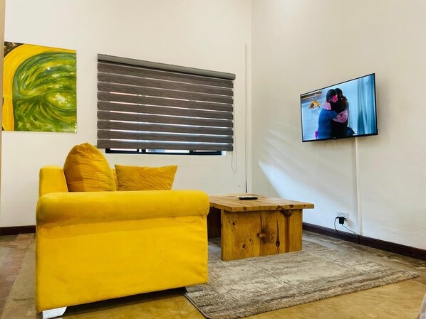 A living room with a yellow sofa, a wooden coffee table, a television mounted on the wall, and a window with blinds. - Amaryllis Residences Mount Pleasant (Blantyre)