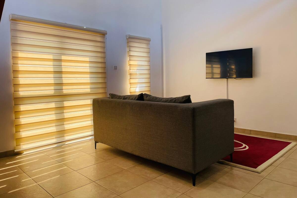 Ein modernes Wohnzimmer mit grauem Sofa, einem an der Wand befestigten Flachbildfernseher und großen Fenstern mit Jalousien.
