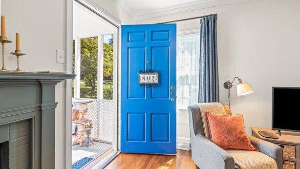 A room with a blue door marked "802", a fireplace, a grey sofa with a throw blanket, and a television on a wooden stand. - 2 Bedroom in Emerywood, High Point, NC (High Point)
