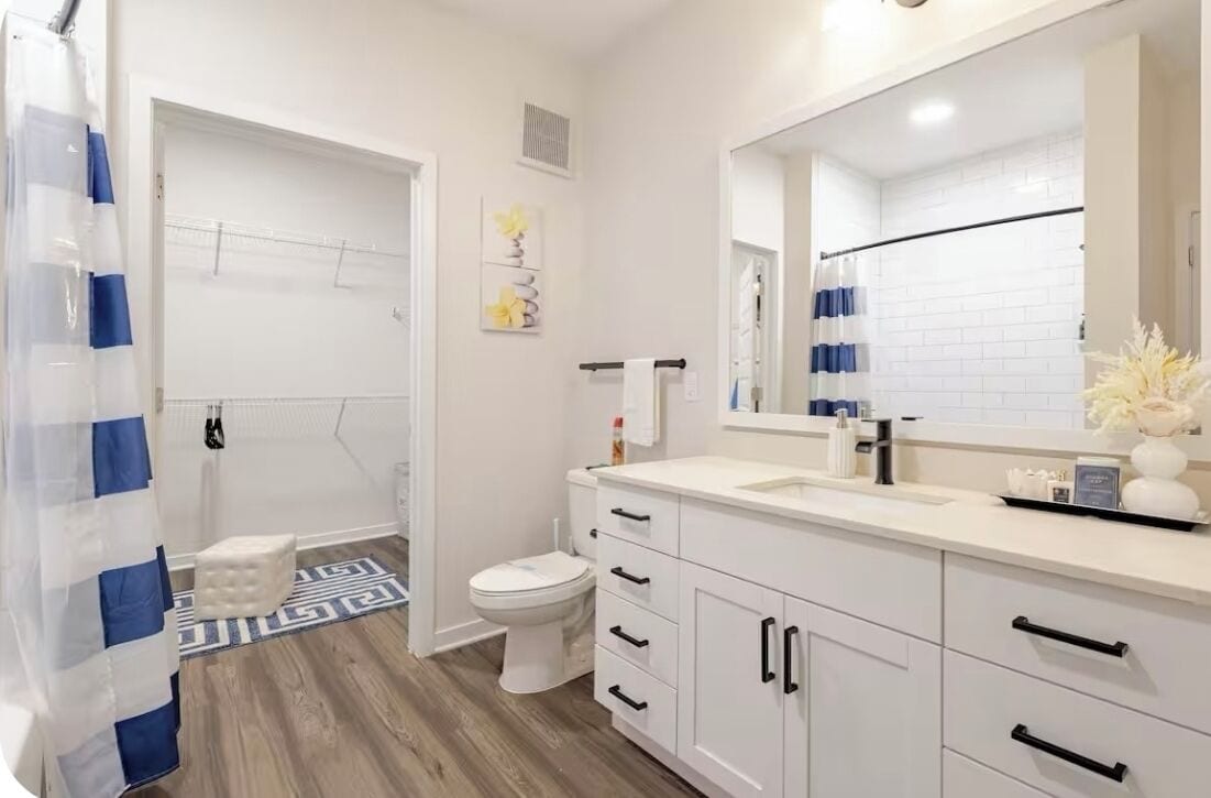 A modern bathroom with a white sink, vanity, and toilet. There is a shower curtain, a small stool, and a decorative vase on the vanity.