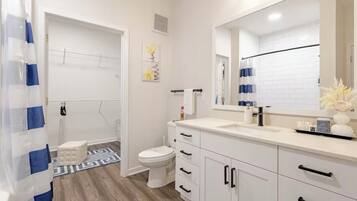 A modern bathroom with a white sink, vanity, and toilet. There is a shower curtain, a small stool, and a decorative vase on the vanity.