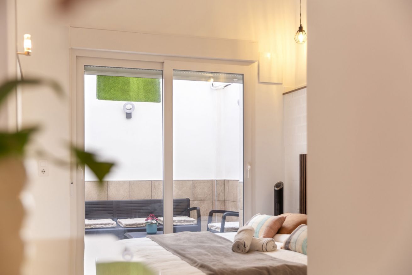Un dormitorio con cama, una mesita de noche, una ventana y una puerta corrediza que da a un espacio exterior con un banco y una planta en maceta.