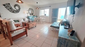 A spacious living room with a sofa, wooden chair, and a coffee table. There is a dining area with a table and chairs, and a large window providing a view of the outdoors. - House on the seafront (La Matanza de Acentejo)