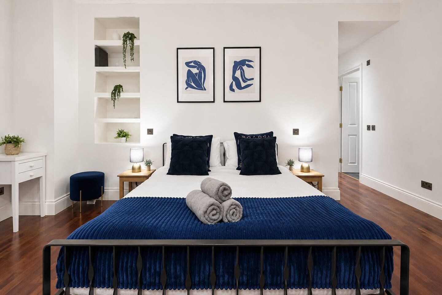 A modern bedroom with a large bed, two bedside tables with lamps, a desk, and a shelving unit with plants.