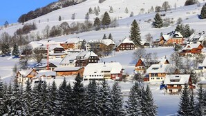 A snow-covered village with traditional wooden houses, a ski lift, and a clear blue sky.