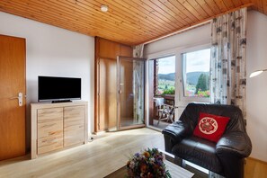 A modern living room with a flat-screen TV, wooden cabinets, a leather sofa with a red pillow, and a glass door leading to an outdoor area with a view of greenery. - Apartment 'Enzian' with Mountain View, Shared Garden and Wi-Fi (Bernau im Schwarzwald)