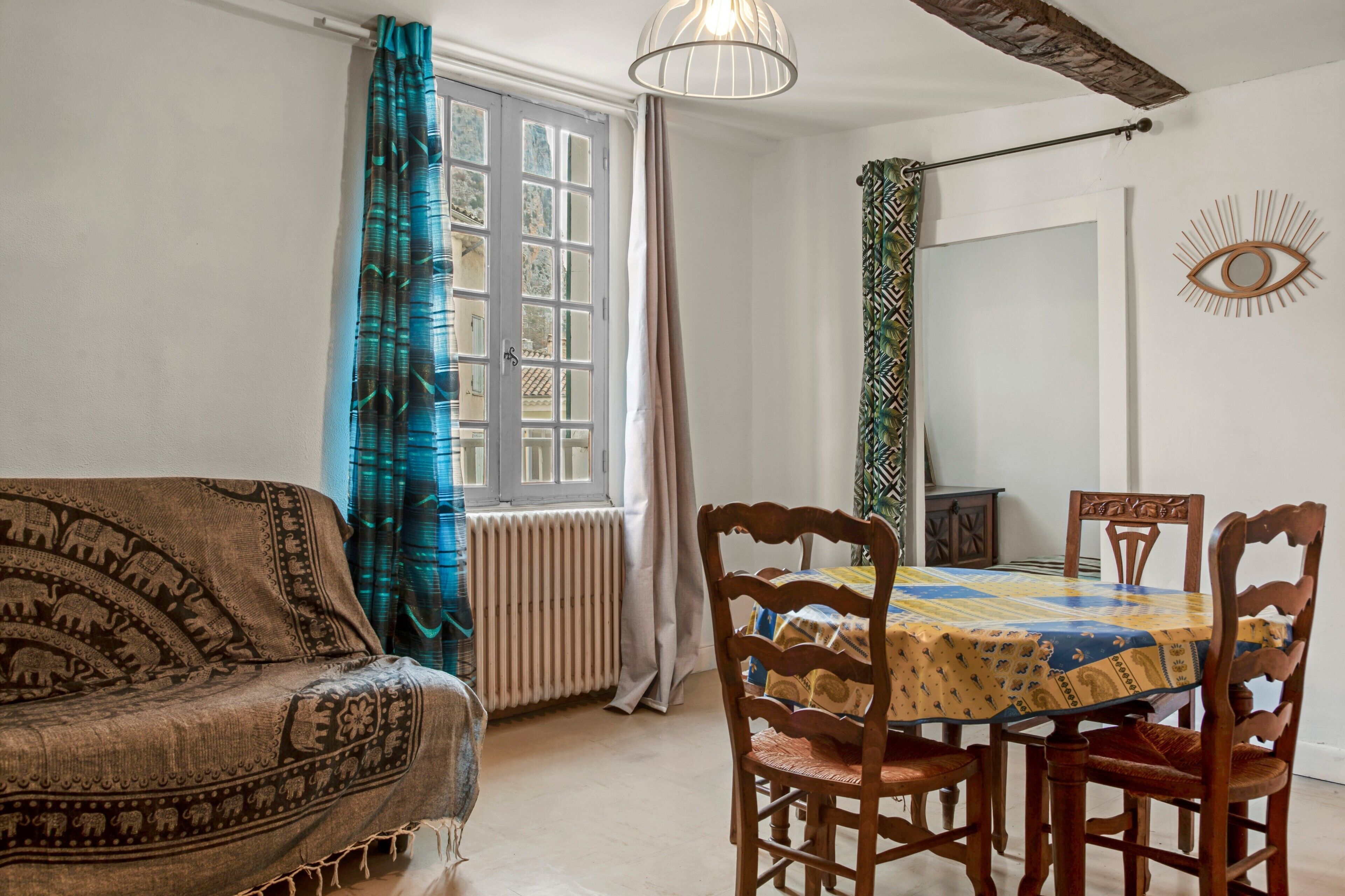A room with a patterned sofa, a dining table with chairs, a radiator, a window with curtains, and a wall-mounted decorative item.