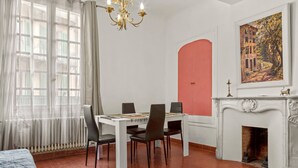 A dining area with a white table, four chairs, a chandelier, a fireplace, and a large window with curtains. - Apartment '1st Floor' with Wi-Fi (Castellane)