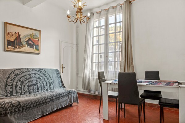 Une pièce comprenant un canapé, une table à manger, des chaises, un lustre, un tableau et une fenêtre avec des rideaux.