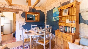 Un interior rústico con paredes de piedra vista, vigas de madera, una mesa de comedor con platos, vasos y un jarrón. Se observa una estantería con libros y objetos decorativos, además de un espejo en la pared.