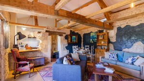 Un interior rústico con vigas de madera a la vista, una pared de piedra, un sofá, una silla, una mesita de centro y una estantería.