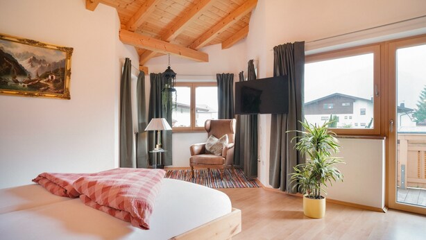 Un dormitorio con cama, una silla, televisor, ventana y una planta.