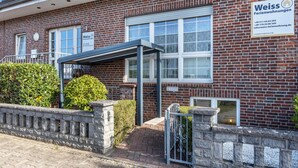 A brick building with a covered entrance, a sign for Weiss Ferienwohnungen, and a stone wall with a gate. - Apartment '3' - Holiday Apartment Weiss (Isernhagen)