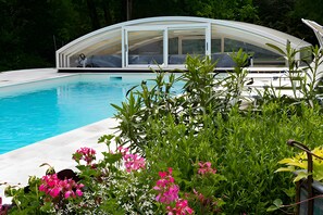 Una piscina amb una zona de solàrium tancada amb vidre, envoltada de vegetació exuberant i plantes florentes.
