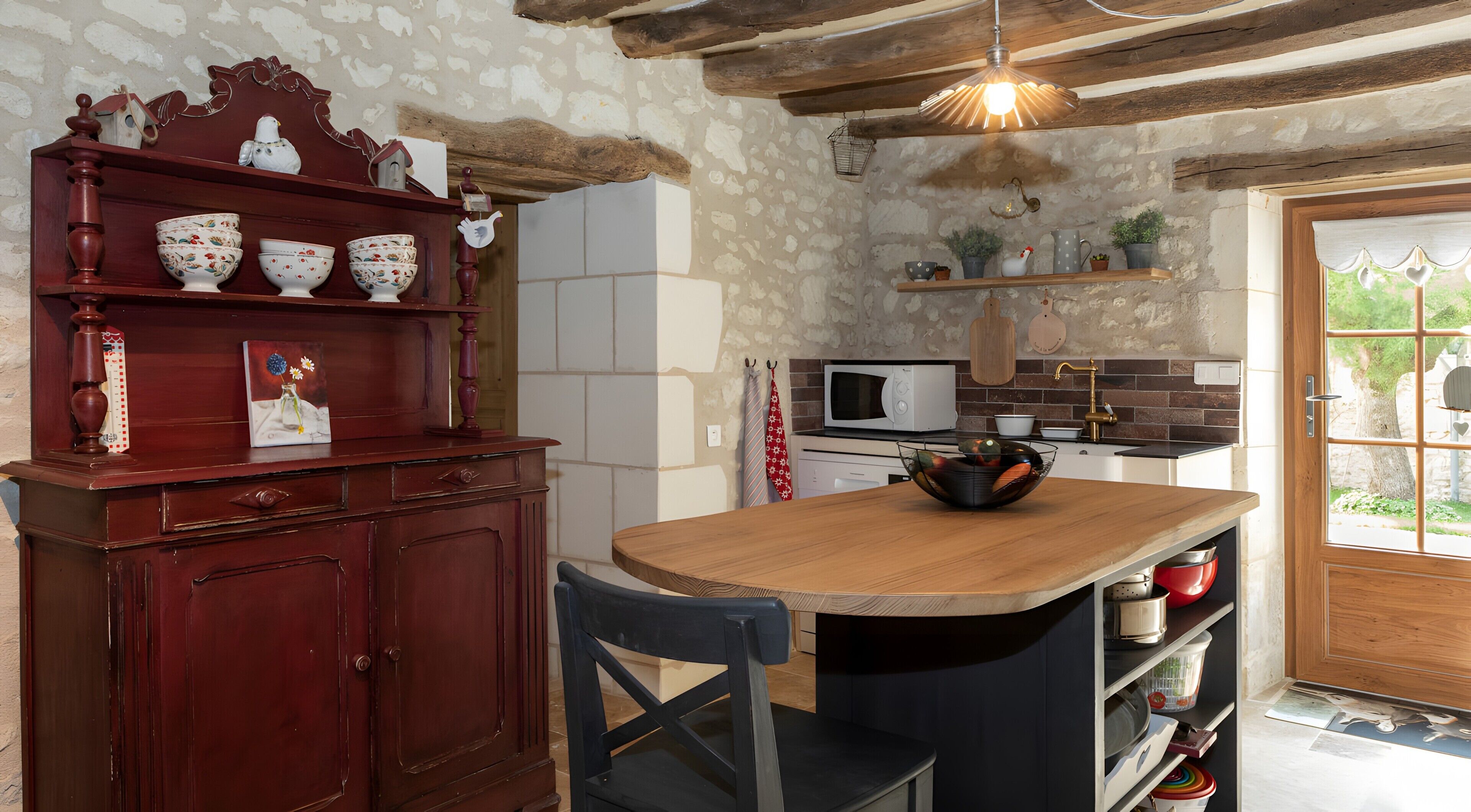 A rustic kitchen with a wooden island, a red cabinet with decorative items, a white microwave, and a view through the window.