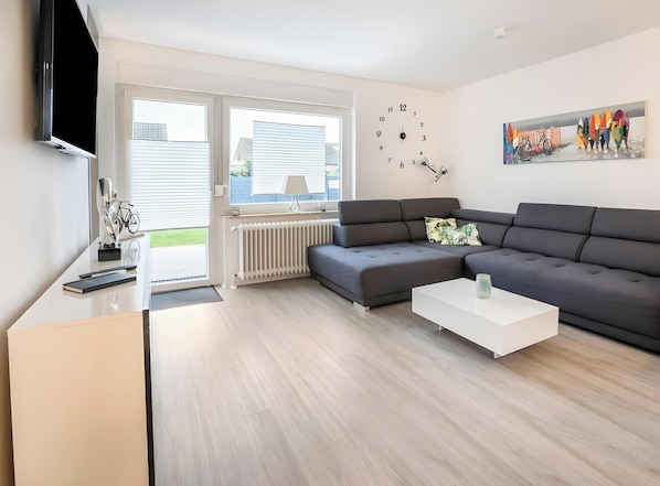 Un salon moderne avec un canapé d’angle, un téléviseur à écran plat, un radiateur et un tableau accroché au mur.