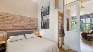 Un dormitorio con pared de ladrillo, una cama con ropa de cama blanca, una mesita de noche con una lámpara, una escalera de madera y una puerta que da a otra habitación.
