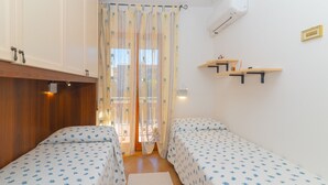 A room with two beds, wooden cabinets, a window with curtains, a wall-mounted air conditioner, and a small shelf with items.