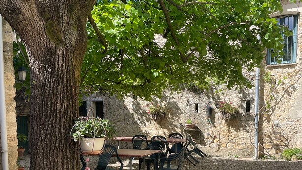 A stone building with a gravel courtyard, outdoor seating, and a large tree providing shade.