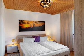 Een slaapkamer met een houten plafond, een bed met witte lakens, een nachtkastje met een lamp en een schilderij van een zonsondergang.