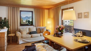 A cozy living room with a wooden ceiling, a sofa with cushions, a dining table with a vase of flowers, and a view of mountains through a window. - Apartment 'Mia' with Mountain View, Balcony and Wi-Fi (Ried im Zillertal)