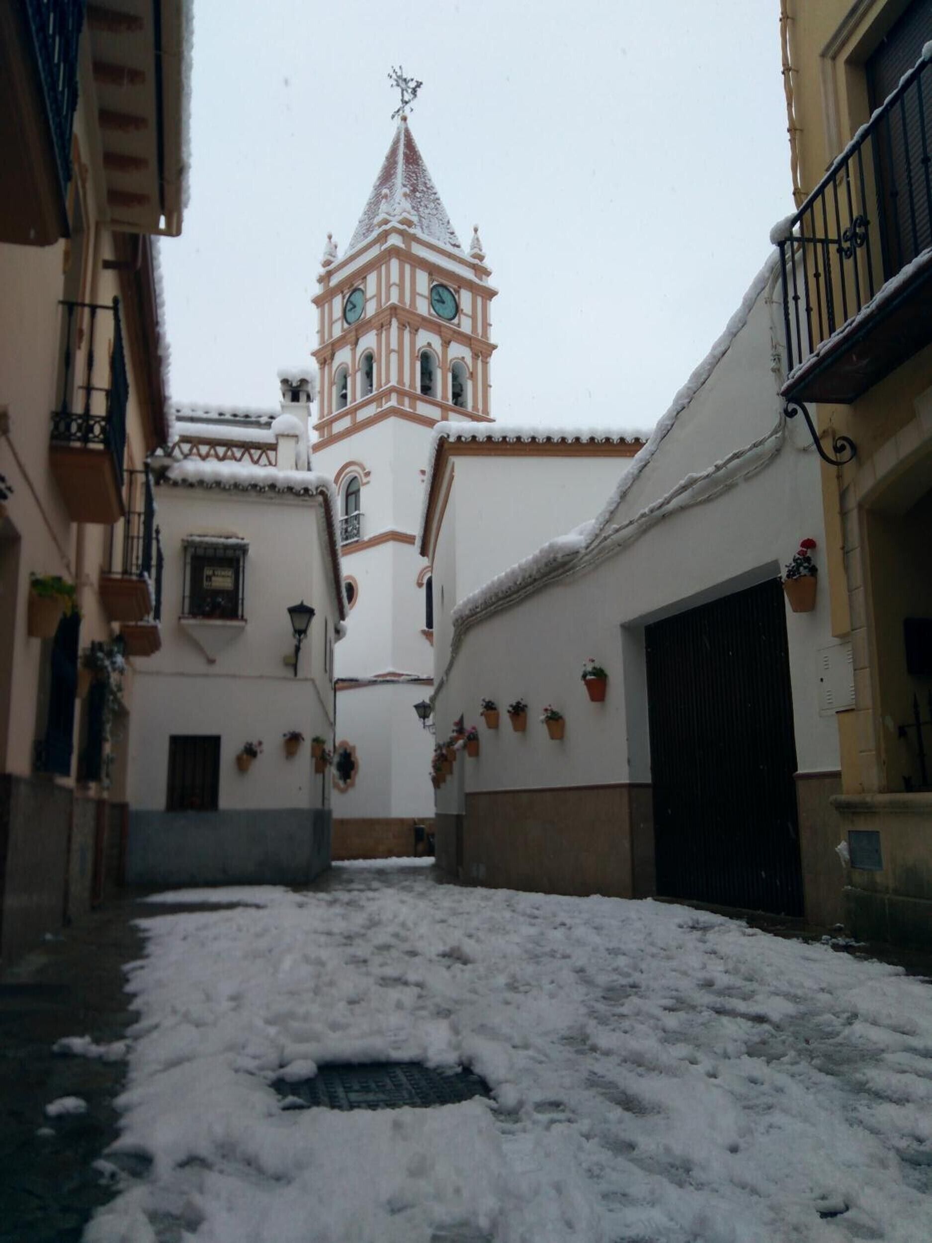 A narrow alleyway with snow-covered ground, a church tower with a clock, and buildings with balconies and potted plants.