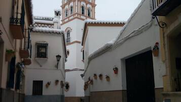 A narrow alleyway with snow-covered ground, a church tower with a clock, and buildings with balconies and potted plants.