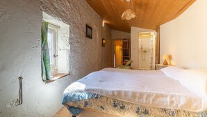 Une chambre avec un plafond en bois, un lit recouvert d’une couvre-lit à motifs floraux, une fenêtre avec des rideaux et une porte donnant sur une salle de bain.