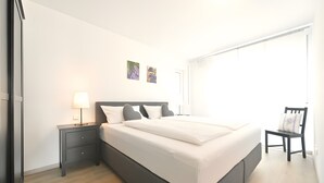 En pænt redt seng med et gråt sengegavl, et natbord med en lampe og en stol med pude.