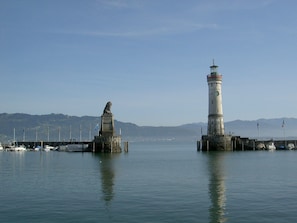 A lighthouse and a statue on a pier with calm water and distant mountains.
