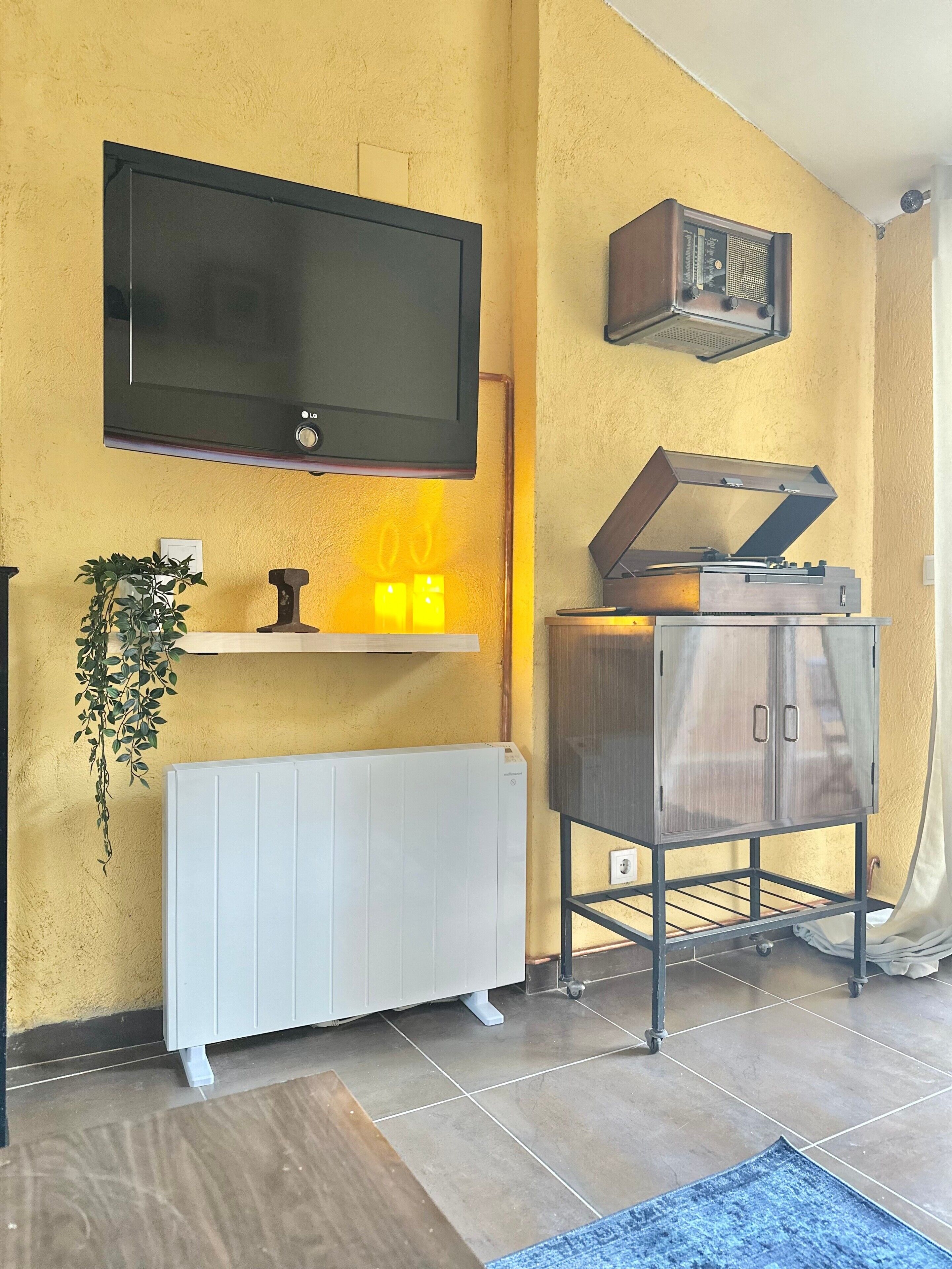 A room with a mounted TV, a radiator, a shelf with candles, a plant, and a record player.